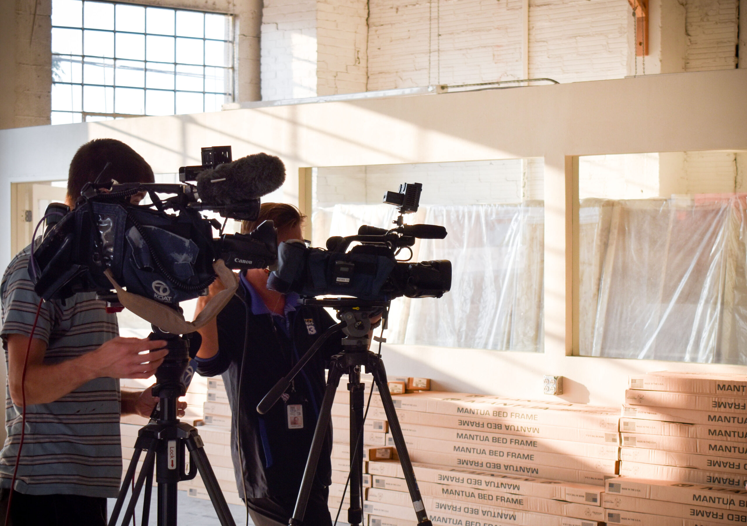A news crew with professional cameras are silhouetted inside a sun filled warehouse.